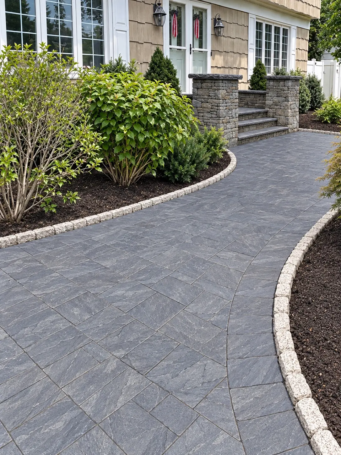 Sweeping gray paver walkway bordered by light stone setts and established plantings